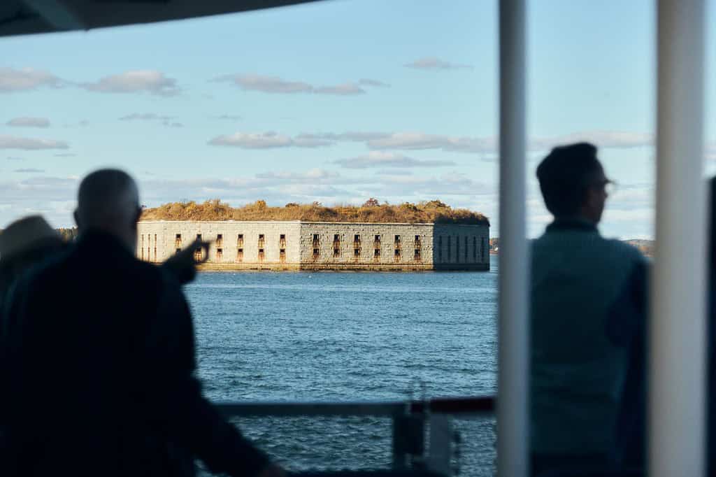 People on a boat look toward a rectangular stone fort on an island surrounded by water, with trees visible on top of the structure under a partly cloudy sky.