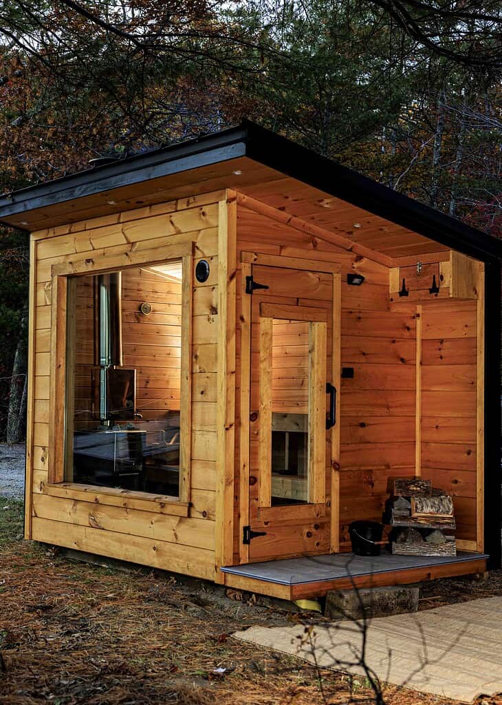 Small wooden cabin with large windows and a slanted roof, set in a wooded area. Firewood is stacked near the entrance, and the interior appears cozy and minimal.
