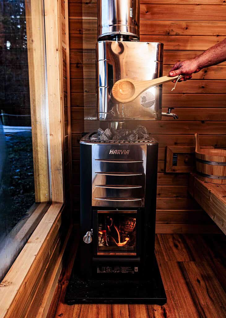 A person pours water with a ladle onto hot stones in a metal sauna stove inside a wooden room.