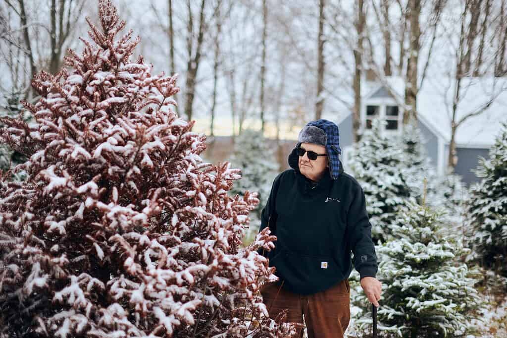 An older man wearing sunglasses and a winter hat stands outdoors near a snow-dusted evergreen tree, holding a cane. Other frosted trees and a small house are in the background.