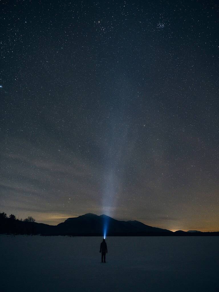 person standing alone in a snowfield at in the night with a headlamp shining into the starry sky