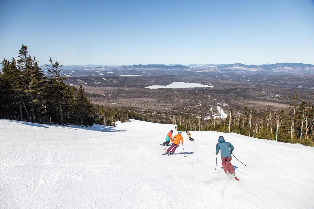 skiers in brightly colored clothing skiing down a slope with blue skies and panoramic valley views below