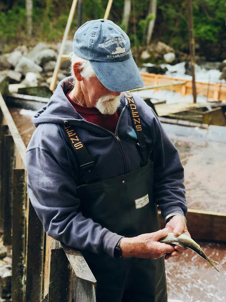 man with a blue hat, white beard, fishing waders and a blue sweatshirt holding a silver fish