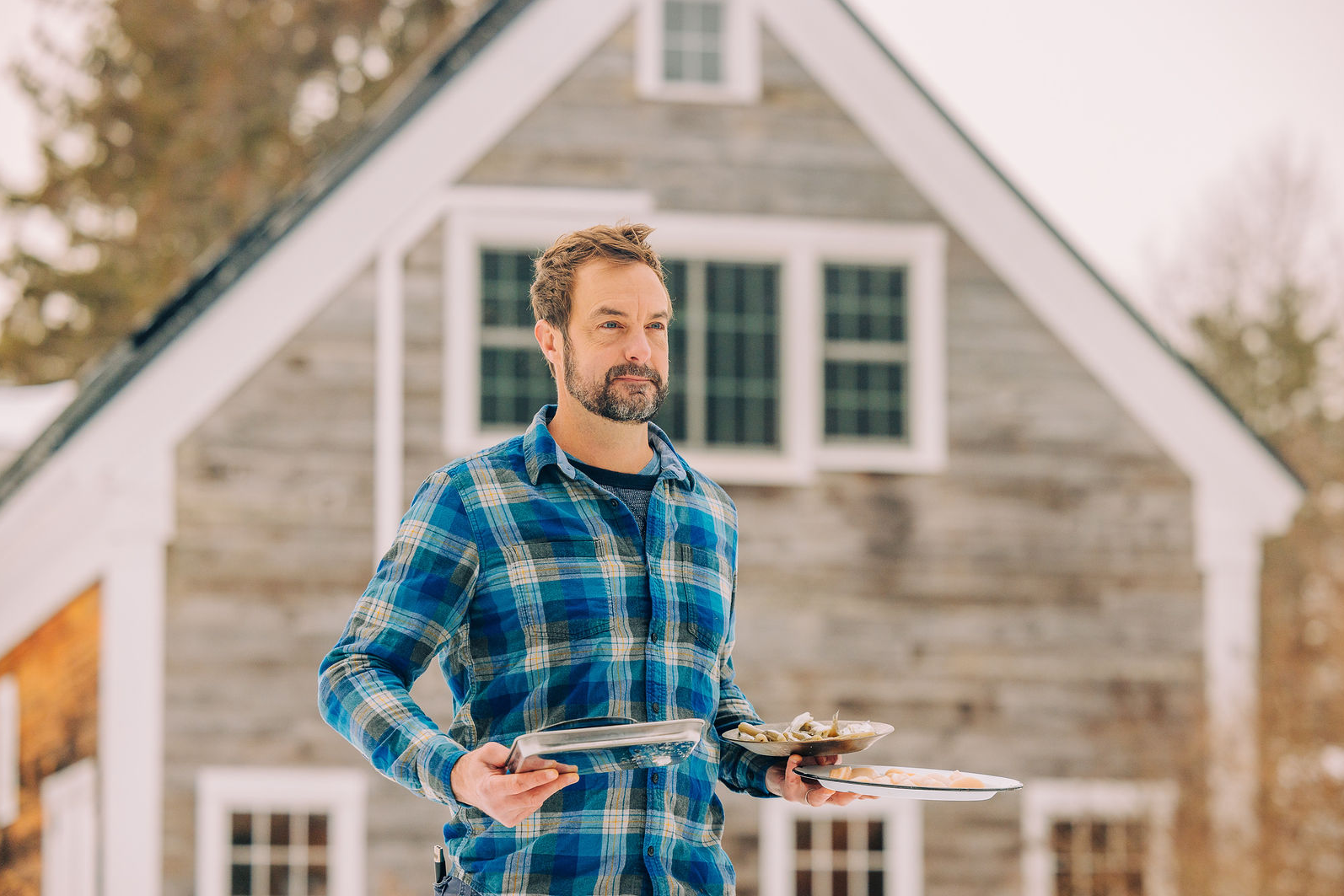A man in a blue plaid shirt stands outside a rustic wooden house, holding two trays with food, one with oysters and one with shrimp. Snow is visible on the roof in the background.