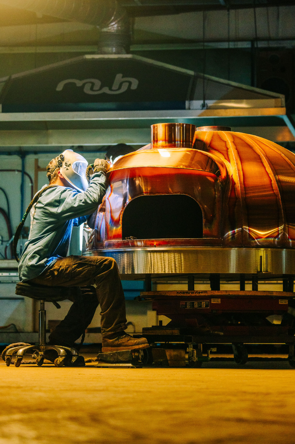 Person wearing protective gear welds a large copper oven in an industrial workshop, sitting on a rolling stool under bright lighting.