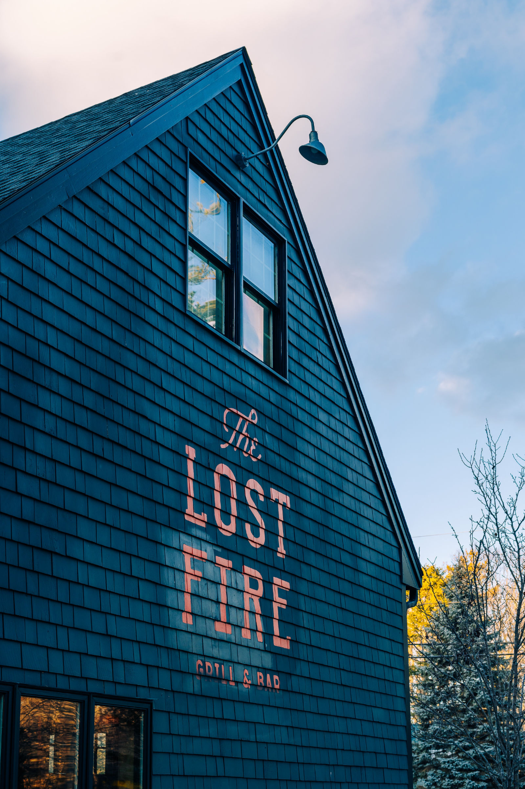 A dark blue shingled building with “The Lost Fire Grill & Bar” painted in large letters on its side, beneath two windows.