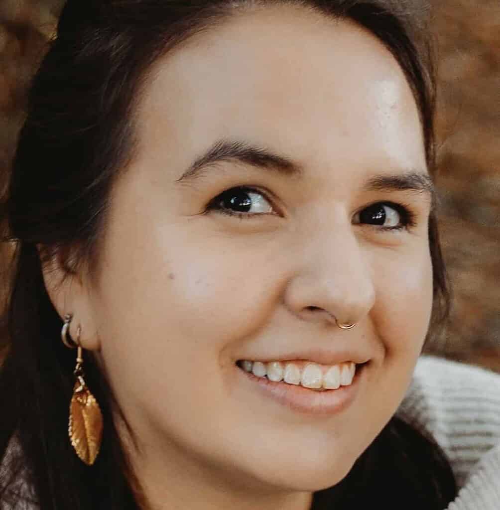 A person with dark hair, wearing a gold leaf earring and a beige sweater, smiles while looking slightly to the side.