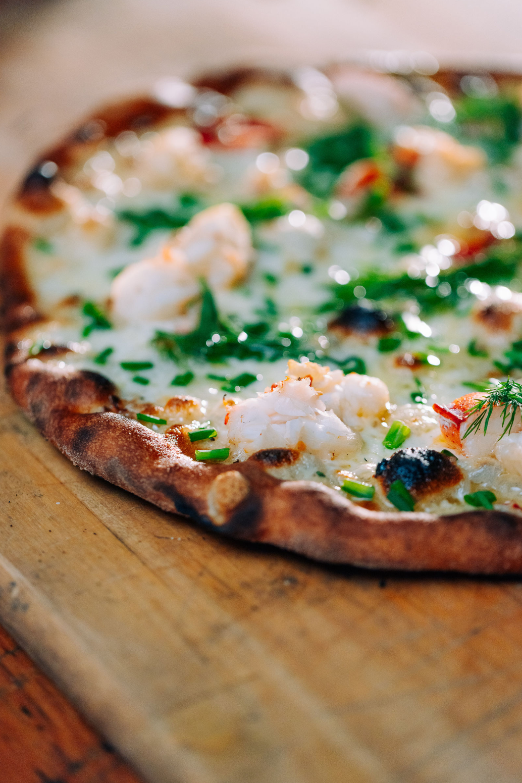 A close-up of a gourmet pizza topped with pieces of lobster, fresh herbs, and melted cheese on a wooden surface.