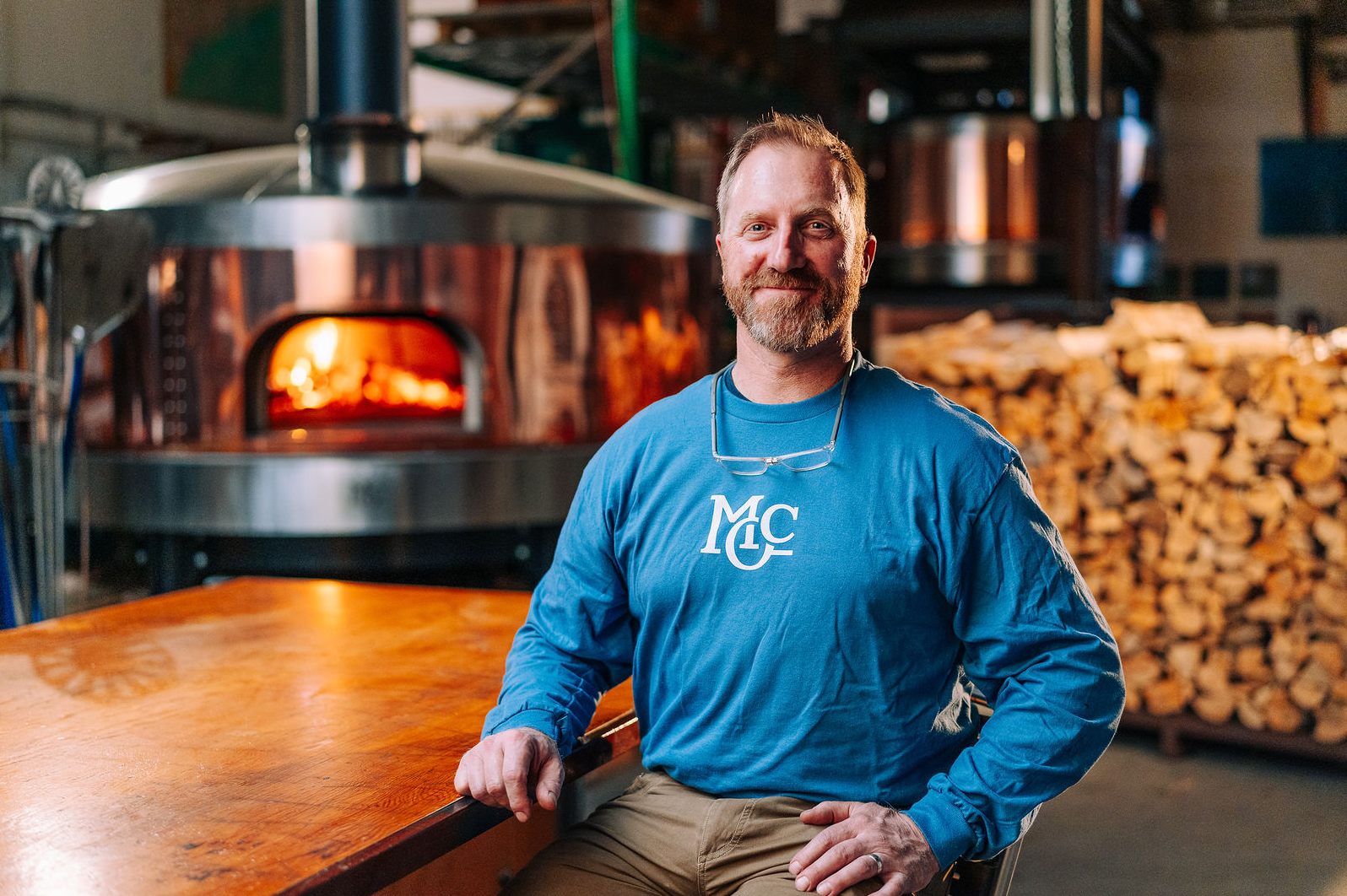 Man in a blue long-sleeved shirt sits at a wooden table in front of a wood-fired oven, with stacked firewood in the background.