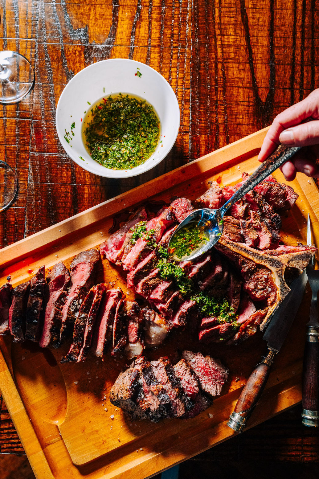 A hand spoons green sauce over sliced grilled steak on a wooden board, with a bowl of sauce and cutlery nearby on a rustic wooden table.