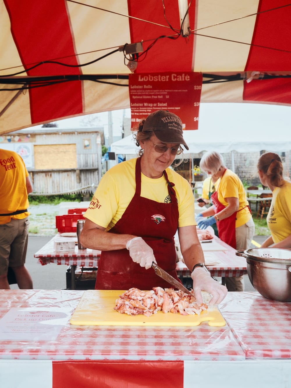 A woman in a red apron and yellow shirt chops lobster meat on a yellow cutting board inside a food tent.
