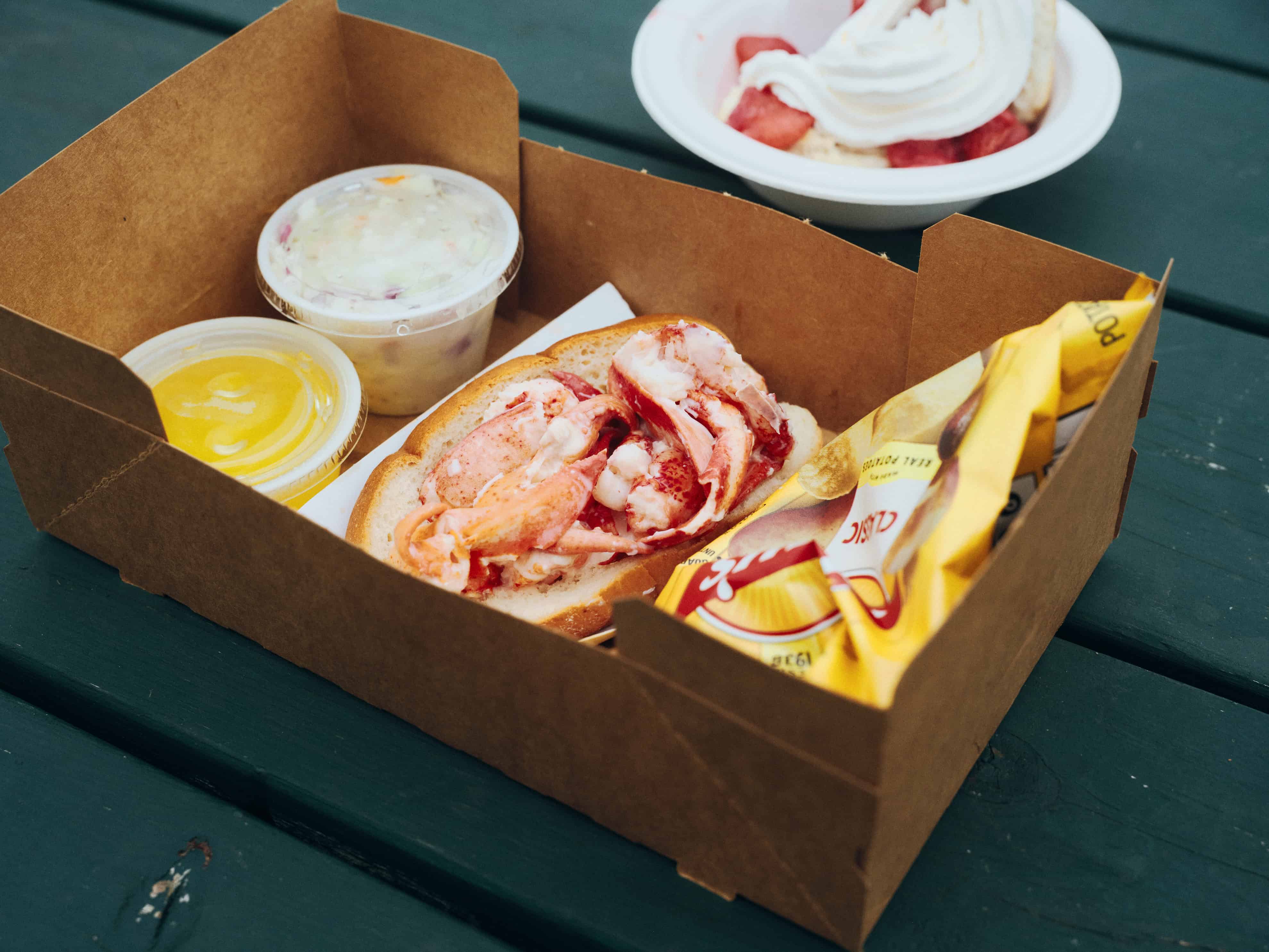 A cardboard takeout box containing a lobster roll, coleslaw, melted butter, and a bag of potato chips, with a side of strawberries and whipped cream.