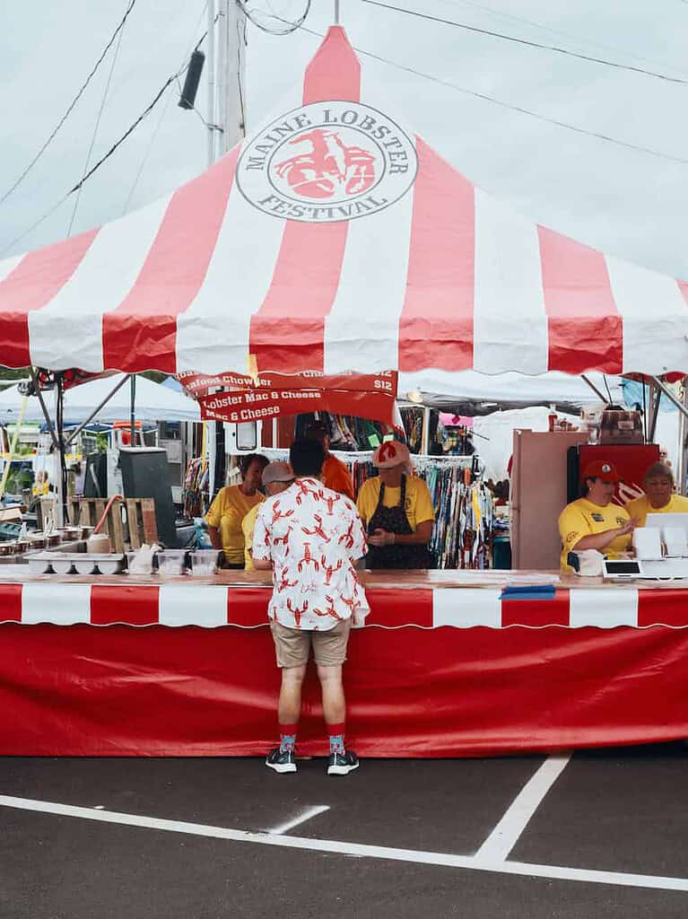 A customer in a lobster-print shirt stands at a food stall under a Maine Lobster Festival tent, while workers in yellow shirts prepare orders behind the counter.