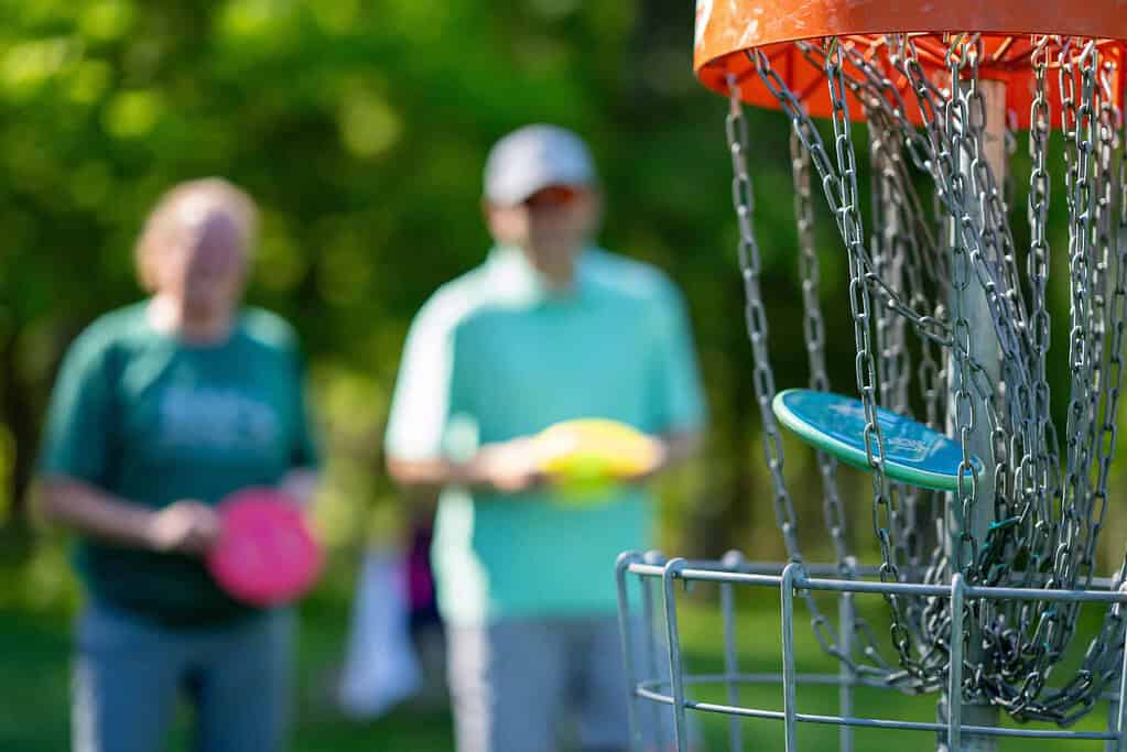 A light blue disc golfer lands in the metal chains of a disc golf basket, with two people blurred in the background.