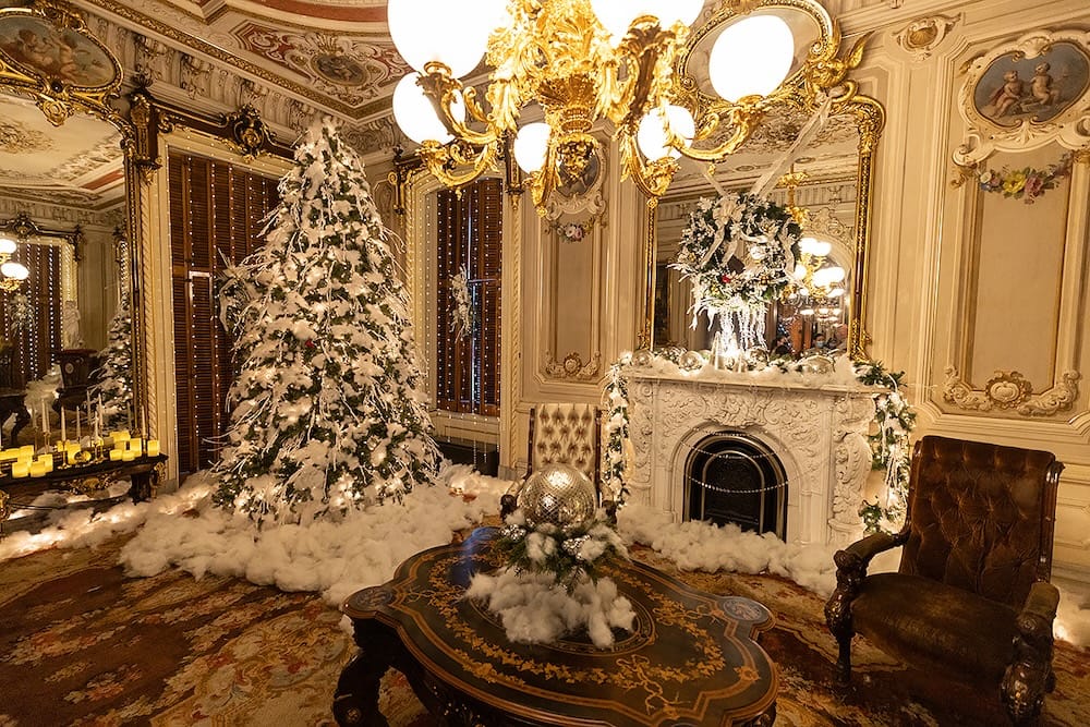 A lavishly decorated room featuring a snow-covered Christmas tree, a fireplace with a festive wreath, a gold chandelier, and ornate antique furniture.