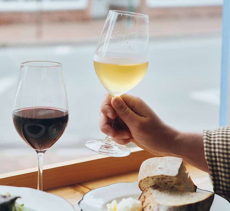 A hand holds up a glass of white wine next to a glass of red wine, bread, and butter on a table by a window.