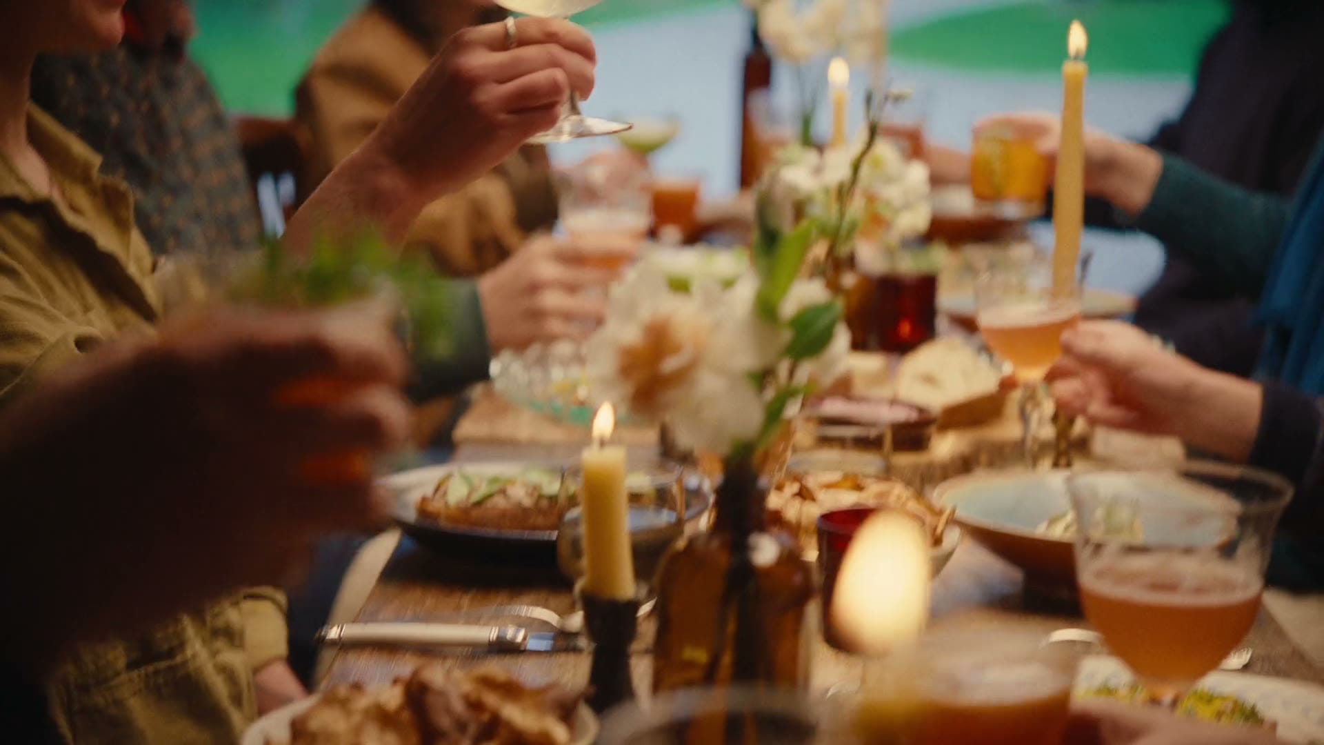 People toast at a dinner table set with candles, flowers, drinks, and food.