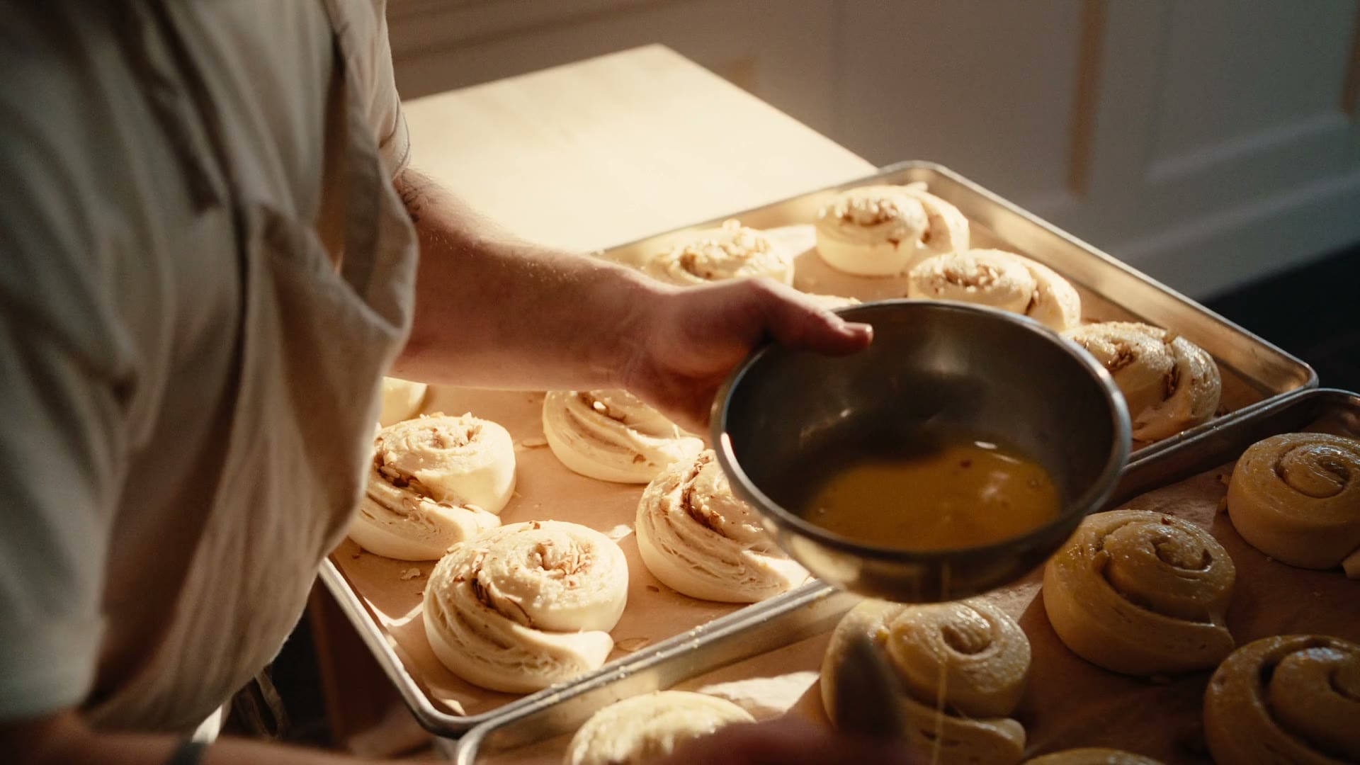 A baker brushes egg wash onto a tray of unbaked cinnamon rolls.